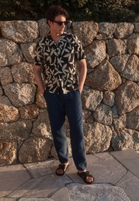 Casual short-sleeve shirt in black with white leaf pattern, paired with loose dark blue pants and brown sandals. Stone wall background.