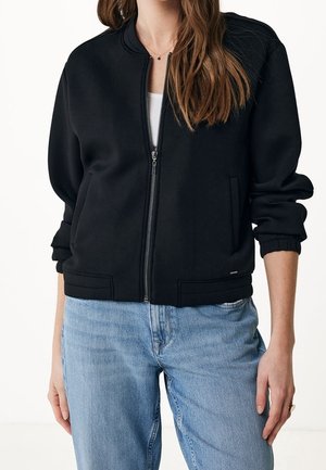 Veste bomber noire zippée avec poignets et ourlet côtelés, portée sur un haut blanc, associée à un jean bleu clair sur une personne debout.