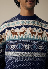 Pulover tricotat cu un model geometric albastru și alb, cu fulgi de zăpadă, cerbi și copaci pe o bază crem, având un guler ribat albastru închis.