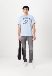 Tricou din bumbac de culoare albastră deschisă cu un imprimeu grafic și blugi din denim gri. Geantă tote neagră ținută în mâna stângă, adidași albi pe picioare.