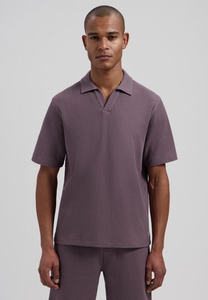 Man met een paarse, kortemouwe, gestructureerde blouse met kraag en bijpassende gestructureerde shorts, staande tegen een effen grijze achtergrond.