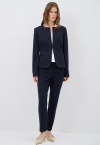 Completo personalizzato blu navy con giacca strutturata e pantaloni coordinati. La giacca presenta un collo arrotondato e una silhouette aderente.