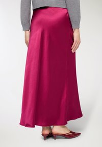 Gonna lunga in raso fucsia con texture liscia. Dettaglio spacco laterale, abbinata a mule con tacco rosso lucido. Maglione grigio lavorato a maglia visibile nella parte superiore.