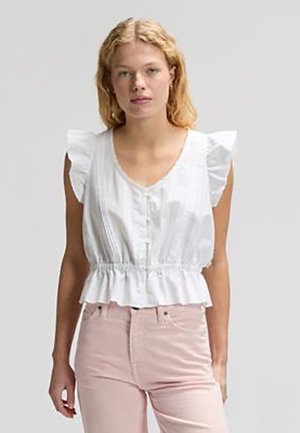 Kobieta o blond włosach, ubrana w białą bezrękawną bluzkę z falbanami na ramionach i jasnoróżowe spodnie, stojąca na tle jednolitego szarego tła.