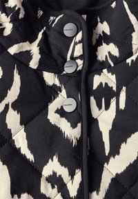 Gestepptes, schwarz-weiß gemustertes Material mit zwei mattschwarzen Knöpfen, versehen mit einem runden Kragen und kontrastierenden Besätzen.