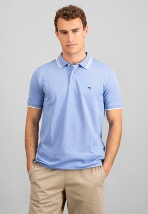 Camisa polo azul clara con ribete blanco en el cuello y las mangas, con un pequeño logo azul oscuro en el pecho. Tejido de algodón suave.