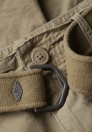 Beżowy pasek z metalową klamrą, teksturowanym paskiem i guzikiem z logo "Superdry", położony na spodniach w podobnym kolorze.