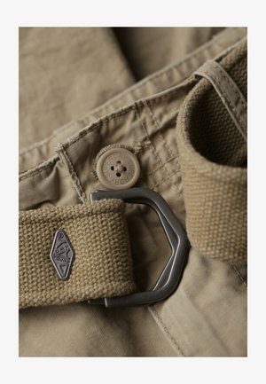 Beige stofbælte med en metalspænde, en tekstureret stofrem og en knap med "Superdry" logo, sat mod bukser i tilsvarende farve.