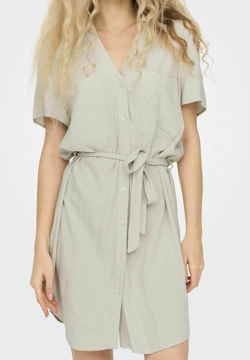 Femme portant une robe boutonnée beige clair à manches courtes avec des poches devant et une ceinture nouée à la taille, debout devant un fond uni.
