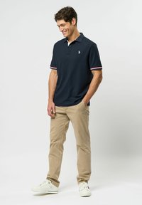 Laivastonsininen poolopaita, jossa on punavalkoraidalliset hihansuut, yhdistettynä vaaleisiin chinoihin ja valkoisiin tennareihin, rinnassa pieni logo.