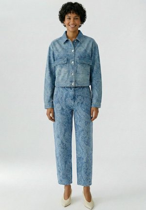 Frau lächelt, steht, trägt eine hellblaue Jeansjacke mit Paisley-Muster und passende hoch taillierte Jeans, dazu weiße spitze High Heels.