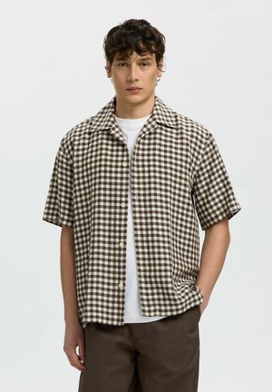 Tânăr purtând o cămașă scurtă cu mâneci și carouri maro și alb peste un tricou alb și pantaloni maro, stând în picioare în fața unui fundal simplu, deschis la culoare.