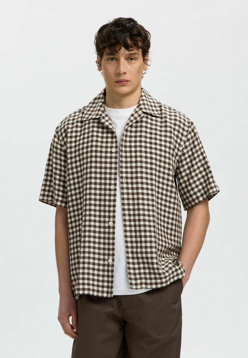 Jeune homme portant une chemise à manches courtes à carreaux marron et blanc sur un t-shirt blanc avec un pantalon marron, debout devant un fond clair uni.