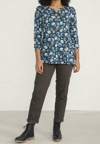 Tunika blu con un motivo a foglie in bianco, marrone e verde acqua, abbinata a pantaloni verde scuro e stivaletti neri. Scollo a V e maniche a tre quarti.
