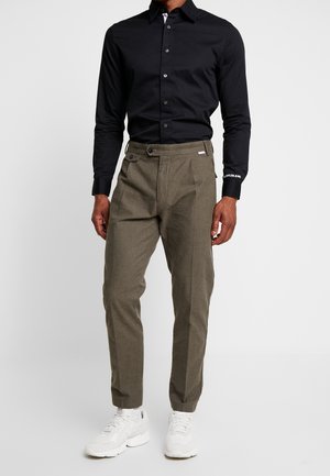 Chemise noire à manches longues, associée à un pantalon plissé vert olive et des baskets blanches. Design simple et couleurs neutres.