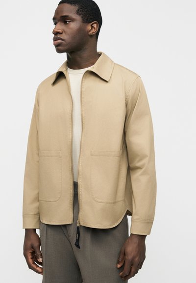 Άνδρας που φοράει μπεζ μπουφάν με φερμουάρ μπροστά και τσεπάκια, πάνω από λευκό πουκάμισο και γκρι παντελόνι, κοιτάζει αριστερά με φόντο ένα απλό φόντο.