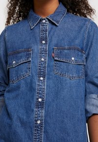 Denimskjorta i medelblått med knäppning fram, två bröstfickor och nedvikbar krage. Utrustad med silverfärgade knappar och uppvikta ärmar.