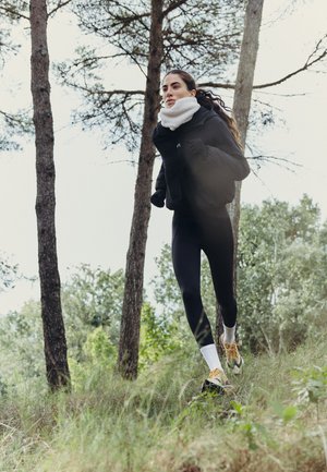 Une femme court dans un sous-bois herbeux, vêtue de vêtements de sport noirs, d’une écharpe blanche et de chaussures de course.