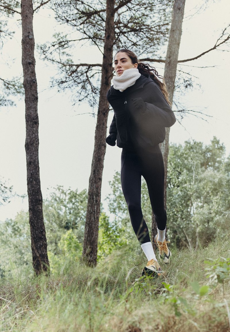 Une femme court dans un sous-bois herbeux, vêtue de vêtements de sport noirs, d’une écharpe blanche et de chaussures de course.