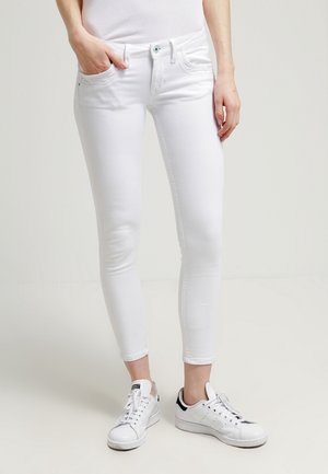 Frau trägt weiße Skinny-Jeans und weiße Sneaker, steht mit einer Hand in der Tasche vor einfachem Hintergrund.