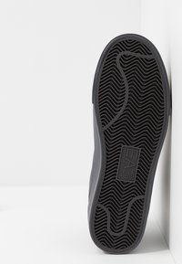 Svart gummisula med ett vågigt texturmönster och en logotyp. Sidan visar en avsmalnande siluett som övergår till en mjuk grå ovandel.