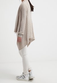 Beige gestrickter Cardigan mit asymmetrischem Saum, getragen zu weißen Leggings und grauen High-Top-Sneakern. Strukturierter Stoff sorgt für visuelles Interesse.