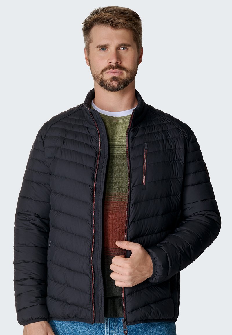 Sort quiltet jakke med høj krave, lynlåslomme foran og kontrastfarvede lynlåse. Bæres over en grøn og rustfarvet stribet sweater.