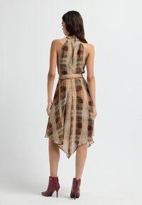Vrouw met lang, donker haar staat met de rug naar ons gekeerd, draagt een mouwloos, bruin gebloemd jurk met een asymmetrische zoom en bordeauxrode enkelhoge laarzen.