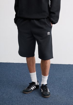 Person trägt schwarze Adidas-Shorts, schwarze Adidas-Sneaker mit weißen Streifen, weiße Socken und einen schwarzen Pullover und steht auf blauem Teppich.