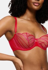 Γυναίκα φορώντας ένα κόκκινο διαφανές σουτιέν με ροζ κεντήματα, πόζαρε με το ένα χέρι στη μέση της σε λευκό φόντο.