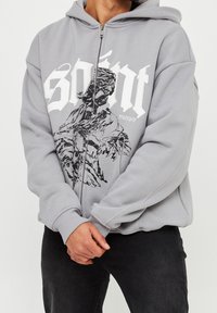 Felpa grigia con zip, caratterizzata da una grande stampa grafica bianca e un design nero sul davanti. Presenta un cappuccio con coulisse e polsini a coste.
