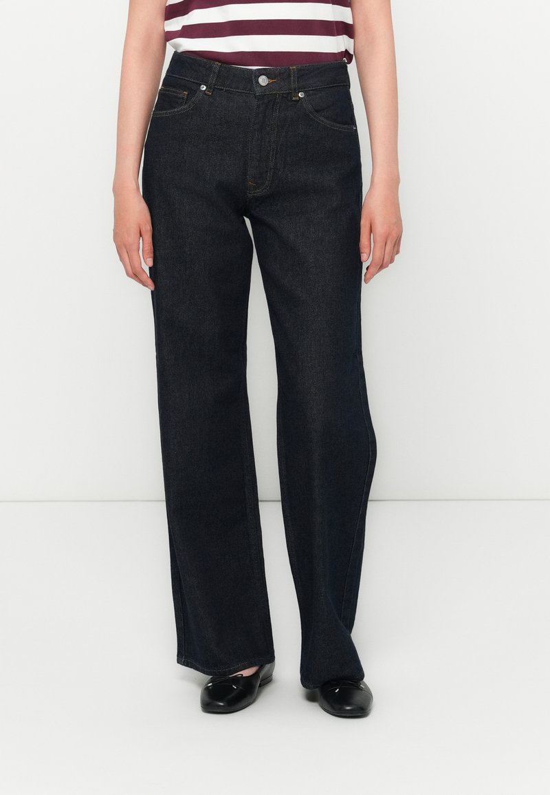 Jean en denim bleu foncé avec une coupe droite, tissu robuste, cinq poches et détails en métal au niveau de la taille.