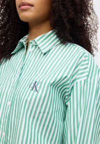 Πράσινο και λευκό ριγέ πουκάμισο από βαμβάκι, με μυτερό γιακά, κλείσιμο με κουμπιά και μικρό λογότυπο στο στήθος.