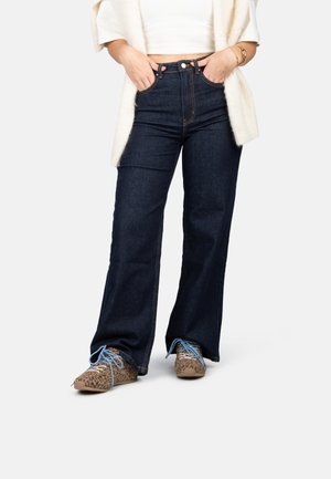 Jean taille haute en denim foncé avec des jambes larges, associés à des chaussures à imprimé léopard marron clair et un top court crème avec un cardigan.