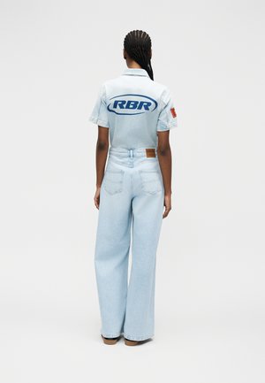 Kvinde med flettet hår iført en lyseblå denim heldragt med "RBR" logo på ryggen, stående mod en ensfarvet hvid baggrund.