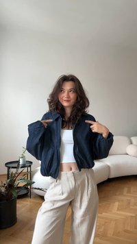 Temno denim bomber jakna z zadrgo, spuščeni rameni in širokimi rokavi, kombinirana z belim skrajšanim topom in bež plisie hlačami.
