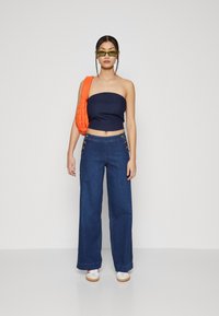 Top sans bretelles marine avec une finition texturée, associé à un pantalon en denim à jambes larges avec des accents de boutons dorés et un sac matelassé orange.