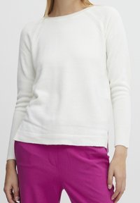 Femme portant un pull blanc à manches longues ajusté et un pantalon rose vif, les mains détendues sur les côtés, contre un fond clair et uni.