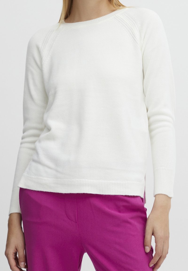 Femme portant un pull blanc à manches longues ajusté et un pantalon rose vif, les mains détendues sur les côtés, contre un fond clair et uni.