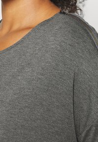 Haut gris à manches courtes avec un col rond, présentant un détail de fermeture éclair le long de l'épaule. Texture douce et coupe décontractée.