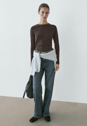 Donna che indossa una maglia a maniche lunghe marrone, un maglione azzurro chiaro legato in vita, jeans blu, scarpe nere e tiene una borsa nera a tracolla.