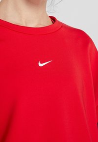 Červené sportovní tričko vyrobené z hladké tkaniny, s kulatým výstřihem a bílým logem Nike na hrudi. Jednoduchý a moderní design.