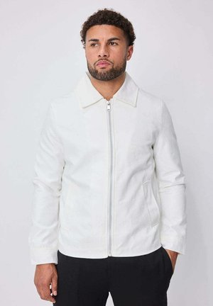 Homme avec barbe et cheveux bouclés portant une veste blanche à fermeture éclair avec col, debout avec une main dans la poche devant un fond clair uni.