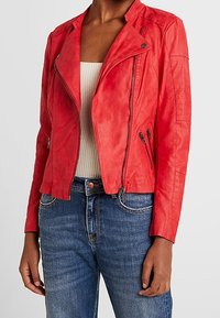 Giacca da motociclista in pelle rossa con zip frontale, tasche laterali e texture trapuntata sulle spalle. Abbinata a un top leggero e jeans di denim.