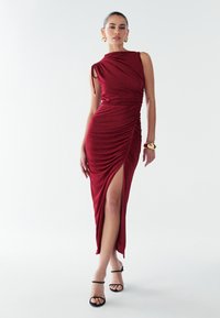 Sarkana sarma kleita ar vienu plecu, rušējumu, augstu sānu šķēlumu, saskaņota ar melnām kurpēm ar siksniņām un zelta apaļām auskarēm.