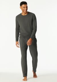 Young man smiling and walking barefoot wearing a dark gray long-sleeve shirt and matching pants against a white background.