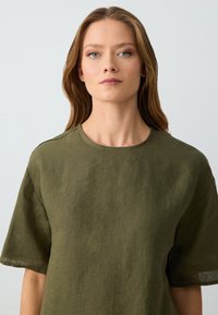 Donna con capelli castano chiaro che indossa una maglietta a maniche corte verde oliva, rivolta verso il fronte su uno sfondo chiaro e semplice.