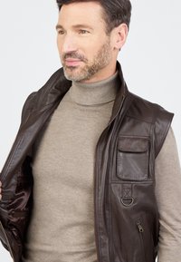 Braune Lederweste mit Reißverschluss, Taschenapplikationen und einer glatten Textur. Trägt man über einem hellbeigen Rollkragenpullover.