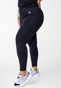 Schwarze Leggings aus dehnbarem Stoff, mit einem Kordelzugbund, seitlichen Taschen und einem kleinen Logo an der Hüfte. Kombiniert mit grauen Sportschuhen.
