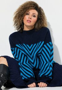 Pull bleu marine avec une texture duveteuse, arborant un motif géométrique en zigzag turquoise. Il possède des poignets côtelés et un col montant.